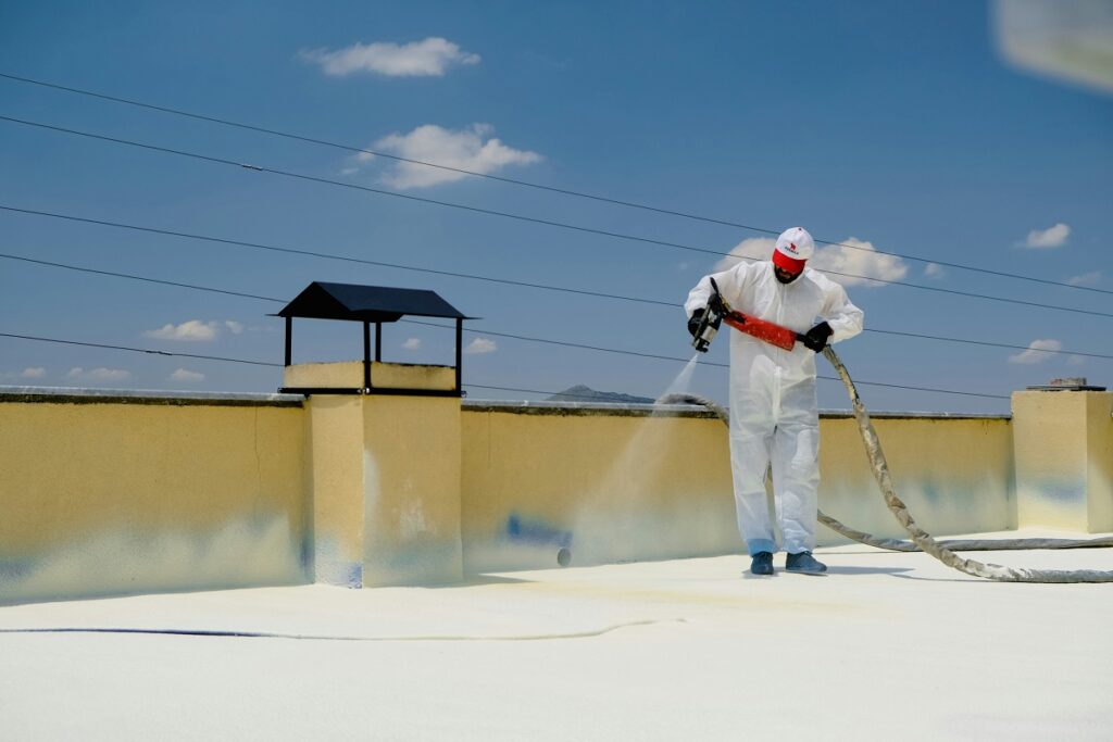 A worker applying white reflective coating to a large flat rooftop under strong sun